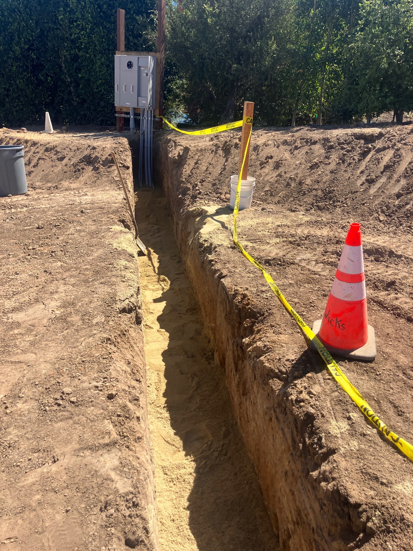 A service trench cut to depth alongside a panel enclosure, caution-taped and ready for conduit before backfill