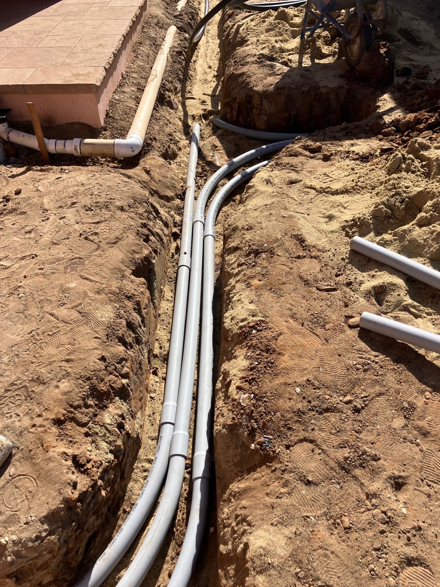Several conduit runs laid side by side down a single trench, sweeping up together toward a wall penetration